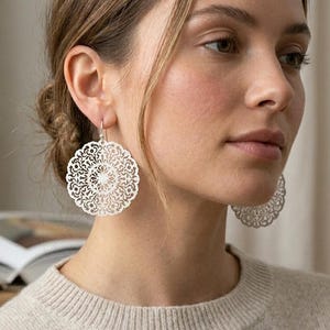 May include: Silver filigree earrings with a circular, openwork design. The earrings are round with intricate patterns, and are hanging from the earlobes. The earrings are a bright silver color and are worn by a person wearing a light-colored sweater.