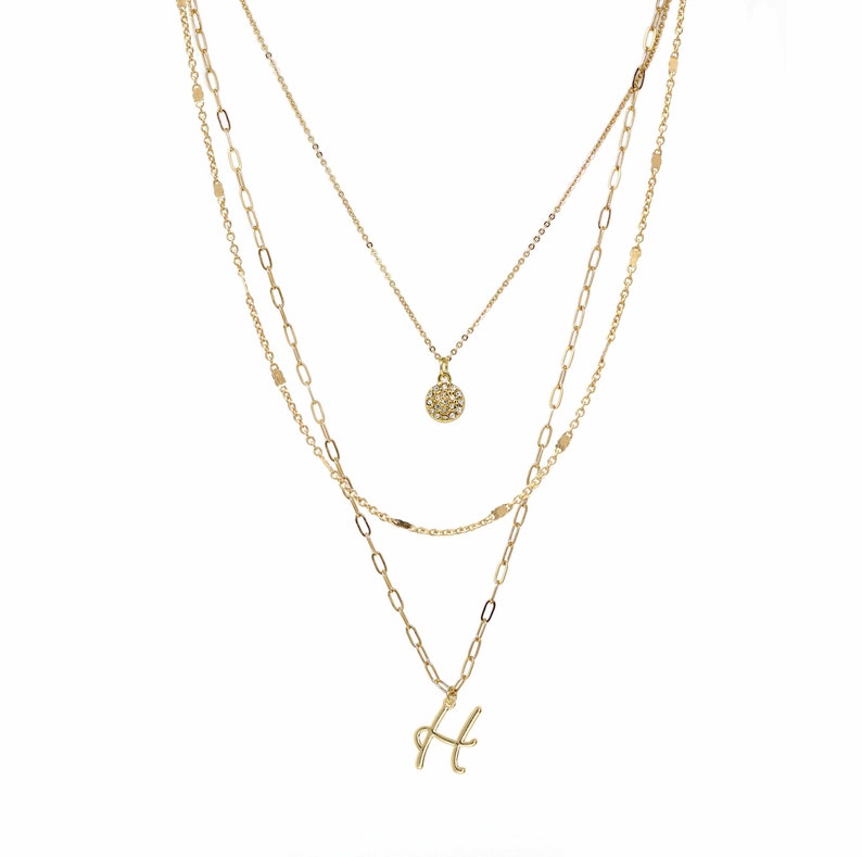 A three-layered gold necklace featuring a script initial charm and a small circle charm paved with stones.