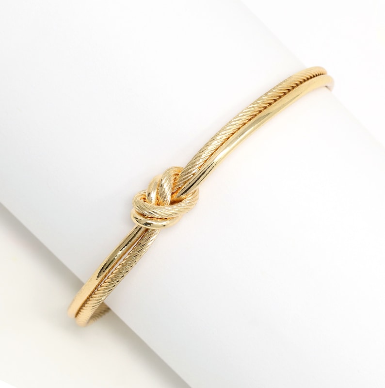 Textured gold knot cuff bracelet displayed on a white cylindrical stand.
