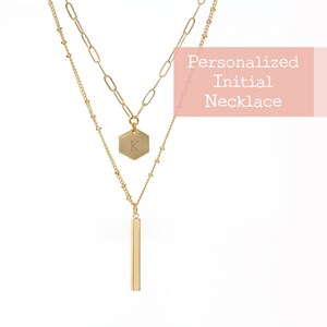May include: Gold layered necklace with a hexagonal pendant engraved with the letter "K" and a long, thin bar pendant.  Personalized Initial Necklace.