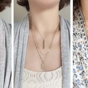 May include: Three gold necklaces with different pendant styles, including a circle, a bar, and a square. The necklaces are layered on a person's neck.