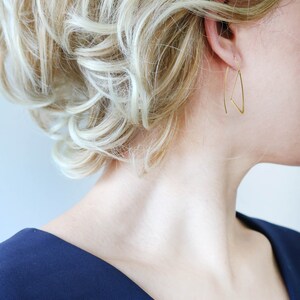K&ouml;nnte beinhalten: Eine Frau mit blonden Haaren tr&auml;gt goldene Dreiecksohrringe. Sie tr&auml;gt ein dunkelblaues Oberteil mit V-Ausschnitt.