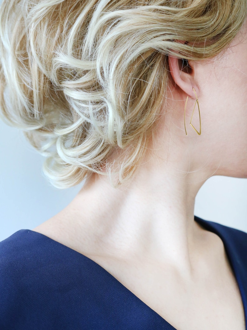 K&ouml;nnte beinhalten: Eine Frau mit blonden Haaren tr&auml;gt goldene Dreiecksohrringe. Sie tr&auml;gt ein dunkelblaues Oberteil mit V-Ausschnitt.