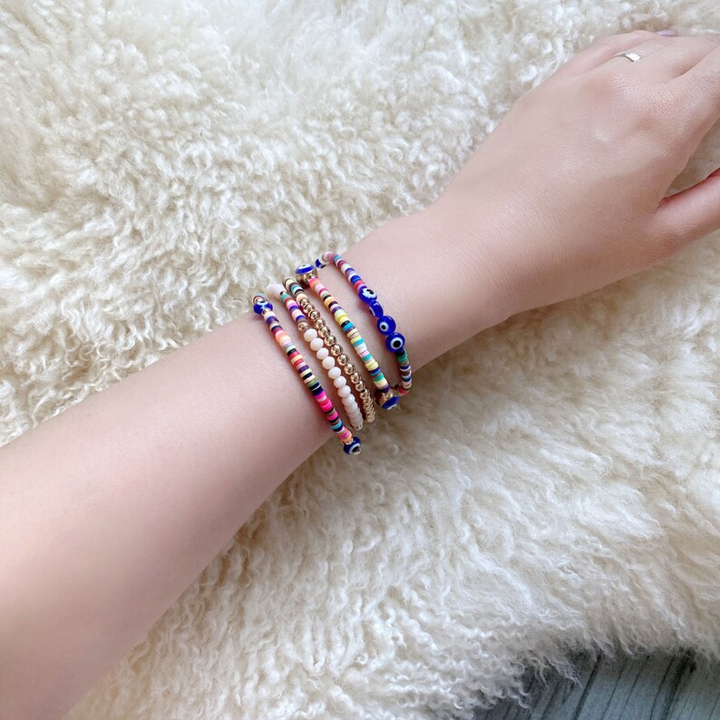May include: A set of six colorful beaded bracelets with blue evil eye charms. The bracelets are stacked on a person's wrist.