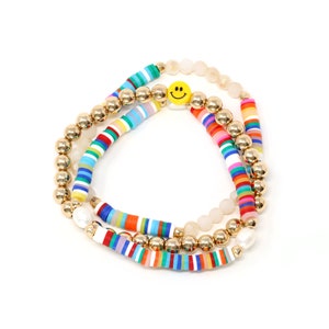 May include: A set of three bracelets featuring colorful striped beads, gold beads, and a single yellow smiley face charm. The bracelets are layered on top of each other.
