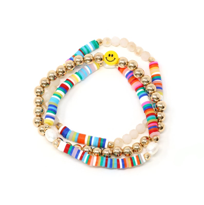 May include: A set of three bracelets featuring colorful striped beads, gold beads, and a single yellow smiley face charm. The bracelets are layered on top of each other.