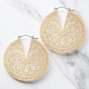 May include: A pair of gold-colored, filigree hoop earrings. Each earring features a circular design with an open center and intricate, symmetrical patterns. The earrings are displayed on a white marble surface.