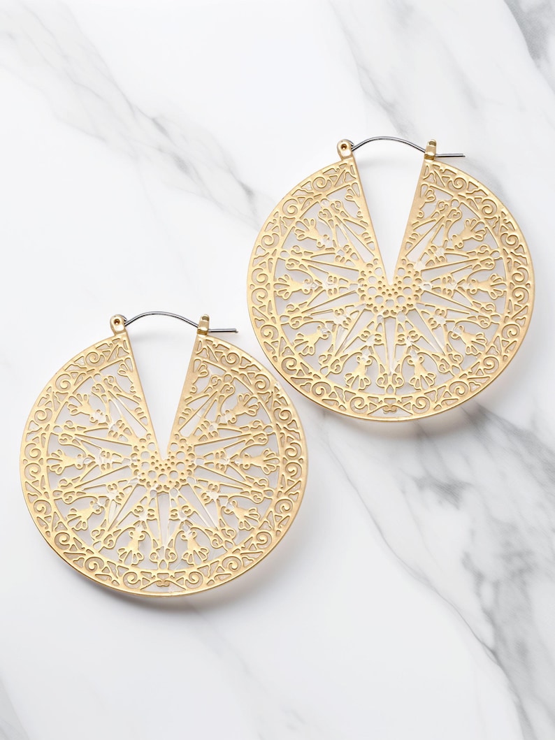 May include: A pair of gold-colored, filigree hoop earrings. Each earring features a circular design with an open center and intricate, symmetrical patterns. The earrings are displayed on a white marble surface.