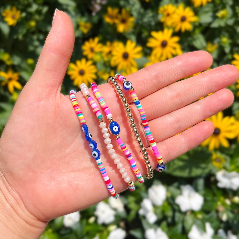 May include: Five colorful beaded bracelets with evil eye charms. The bracelets are stacked on a hand and feature pink, blue, white, yellow, and green beads.