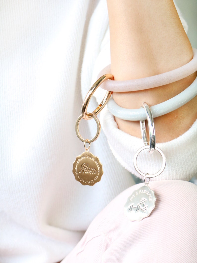 May include: Two gold and silver keychains with charms. The gold charm has the word "Mom" and the text "Philosophers have lost much sleep over the question of whether we are getting better or worse." The silver charm has the text "Fit & Fabulin" and a dumbbell graphic.