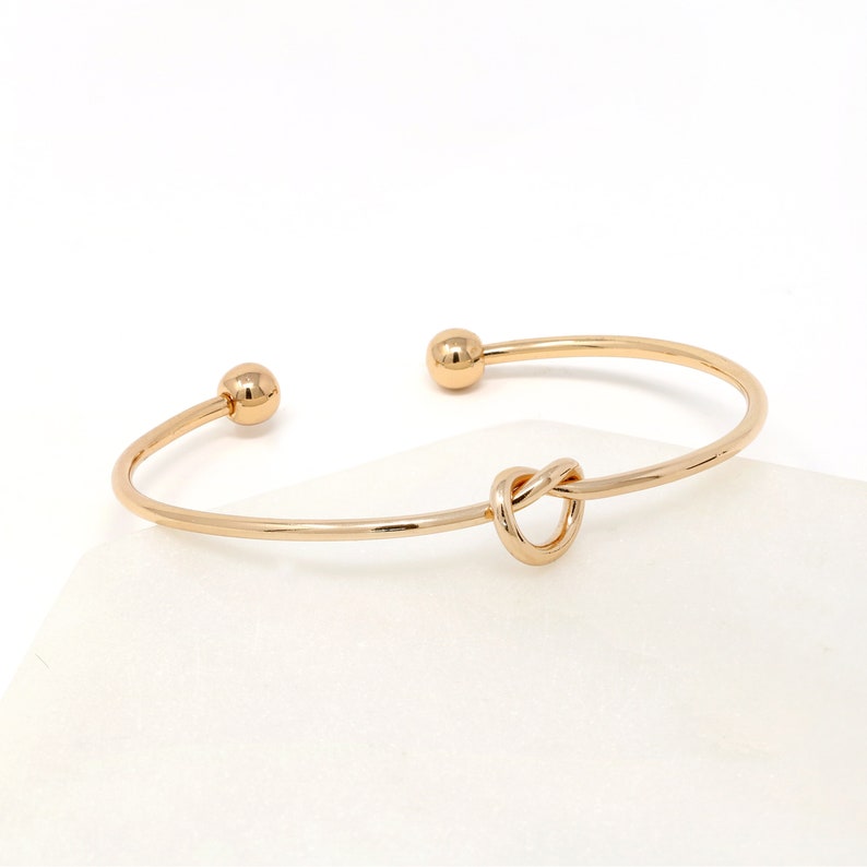 Thin gold open cuff bracelet with a loop knot front and ball ends, on a white marble surface.