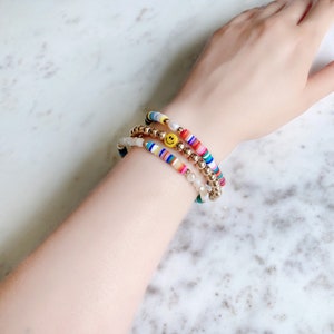 May include: Three bracelets stacked on a wrist. The top bracelet is a gold chain with a yellow smiley face charm. The middle bracelet is a white beaded bracelet with gold beads. The bottom bracelet is a multi-colored striped bracelet with gold beads.