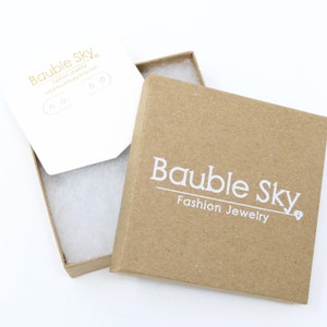 Puede incluir: Una caja de cart&oacute;n marr&oacute;n con una etiqueta blanca que dice "Bauble Sky Fashion Jewelry". La caja est&aacute; abierta y el interior est&aacute; forrado con papel de seda blanco.