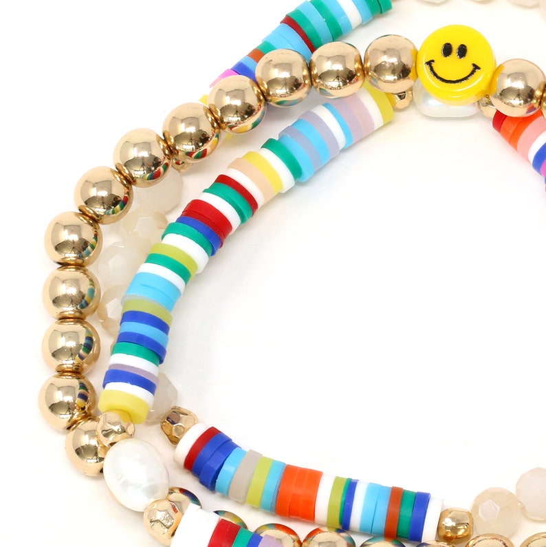 May include: A set of three bracelets featuring colorful striped beads, gold beads, and a white pearl bead with a yellow smiley face charm.