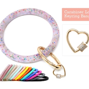 May include: A white, glitter-speckled bangle with a gold-coloured heart-shaped carabiner. The image also shows a selection of colourful bangles and the text "Carabiner Lock Keyring Bangle."