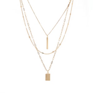 May include: A gold layered necklace with two pendants. The top pendant is a long, thin bar and the bottom pendant is a square.