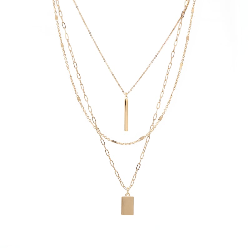 May include: A gold layered necklace with two pendants. The top pendant is a long, thin bar and the bottom pendant is a square.