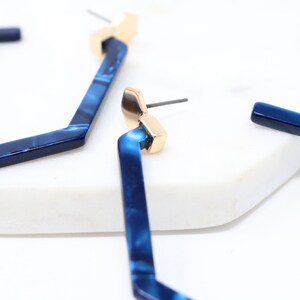 May include: A pair of blue and gold geometric hoop earrings. The earrings are made of a blue marbled material and have a gold-toned post.