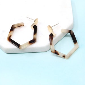 May include: A pair of geometric hoop earrings with a tortoiseshell pattern in shades of brown and cream. The earrings have a gold-colored post and are displayed on a white surface against a light blue background.