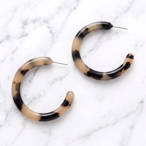 May include: A pair of hoop earrings featuring a smooth, rounded profile with a translucent beige and dark brown tortoiseshell pattern. Each earring is constructed as an open circle with a thin metal post extending from the top edge for attachment.