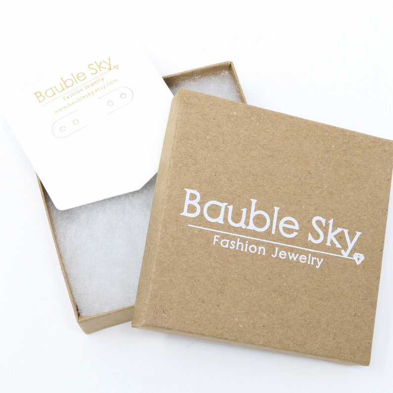 Puede incluir: Caja de joyer&iacute;a de cart&oacute;n marr&oacute;n abierta con las palabras "Bauble Sky Fashion Jewelry" impresas en blanco. Dentro hay una tarjeta blanca para exhibir pendientes, con el logotipo y la direcci&oacute;n del sitio web de Bauble Sky.