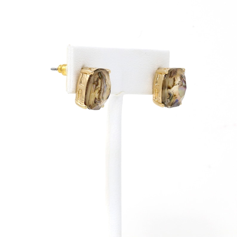 Puede incluir: Un par de pendientes de bot&oacute;n dorados. Cada pendiente presenta una piedra rectangular iridiscente engastada en un marco dorado. Los pendientes se exhiben en un soporte blanco para pendientes sobre un fondo blanco.