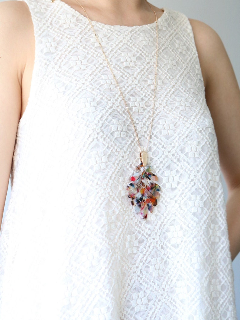 May include: A gold chain necklace with a multi-colored leaf pendant. The pendant is made of clear plastic with red, orange, yellow, green, blue, and purple accents. The necklace is worn by a person wearing a white lace top.