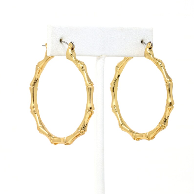 May include: A pair of gold hoop earrings with a bamboo-like design. The earrings are displayed on a white stand.