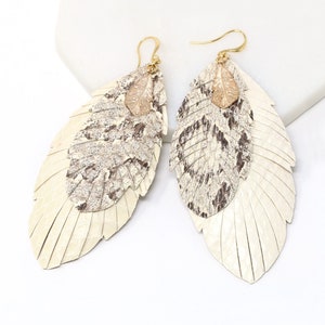 Op de afbeelding: Een paar veren oorbellen met een gelaagd ontwerp. De bovenste laag is een bruin en wit gepatroneerde veer, en de onderste laag is een witte veer. De oorbellen hebben goudkleurige accenten.