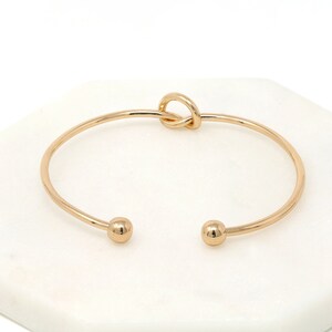Top view of the thin gold cuff bracelet with ball ends and a loop knot, on a white marble tile.