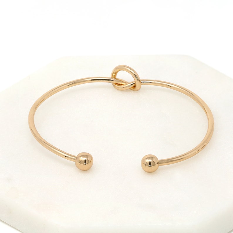 Top view of the thin gold cuff bracelet with ball ends and a loop knot, on a white marble tile.