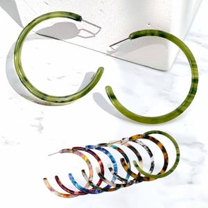 May include: A collection of hoop earrings featuring a variety of colors and patterns. Two large olive green translucent hoops are positioned at the top, while a stack of smaller hoops in assorted marbled patterns including blue, red, and brown sits below.