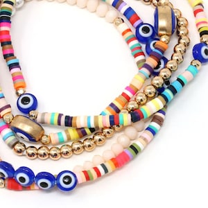 May include: Multicolored beaded bracelets with gold beads and blue evil eye charms. The bracelets are layered and feature a variety of colors, including pink, red, yellow, green, blue, and purple.