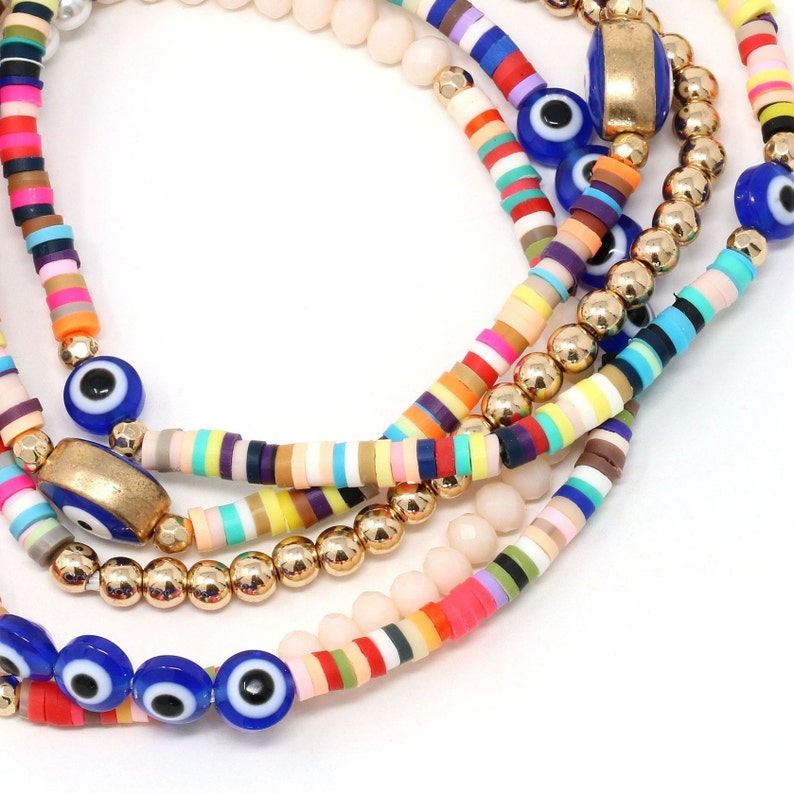 May include: Multicolored beaded bracelets with gold beads and blue evil eye charms. The bracelets are layered and feature a variety of colors, including pink, red, yellow, green, blue, and purple.