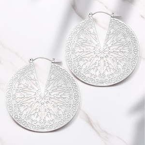May include: Two silver-colored, open-hoop earrings. Each earring is a large, circular design with an intricate, openwork pattern. The pattern features a central starburst design surrounded by swirling, floral-like details. The earrings are displayed on a white marble surface.