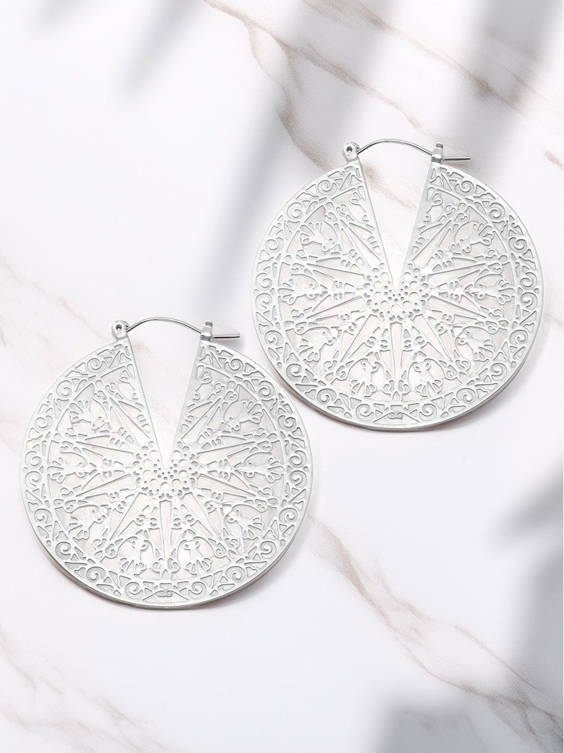 May include: Two silver-colored, open-hoop earrings. Each earring is a large, circular design with an intricate, openwork pattern. The pattern features a central starburst design surrounded by swirling, floral-like details. The earrings are displayed on a white marble surface.