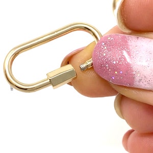 May include: A gold-toned metal clasp with a rounded oval shape. The clasp is open and held by a finger with pink and white nail polish.