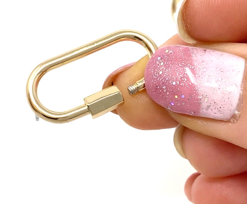 May include: A gold-toned metal clasp with a rounded oval shape. The clasp is open and held by a finger with pink and white nail polish.
