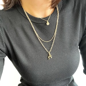 Puede incluir: Una mujer lleva una blusa negra de canal&eacute; con tres collares de oro. Los collares tienen un peque&ntilde;o colgante redondo, un colgante de eslab&oacute;n de cadena y un colgante con la letra 'A'.