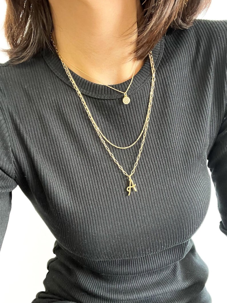 Puede incluir: Una mujer lleva una blusa negra de canal&eacute; con tres collares de oro. Los collares tienen un peque&ntilde;o colgante redondo, un colgante de eslab&oacute;n de cadena y un colgante con la letra 'A'.