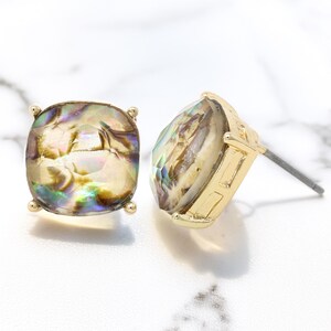Puede incluir: Dos pendientes de bot&oacute;n dorados con piedras cuadradas iridiscentes. Las piedras muestran una mezcla de colores, incluyendo oro, verde y p&uacute;rpura. Cada pendiente tiene un poste para orejas perforadas y peque&ntilde;os detalles dorados en las esquinas.