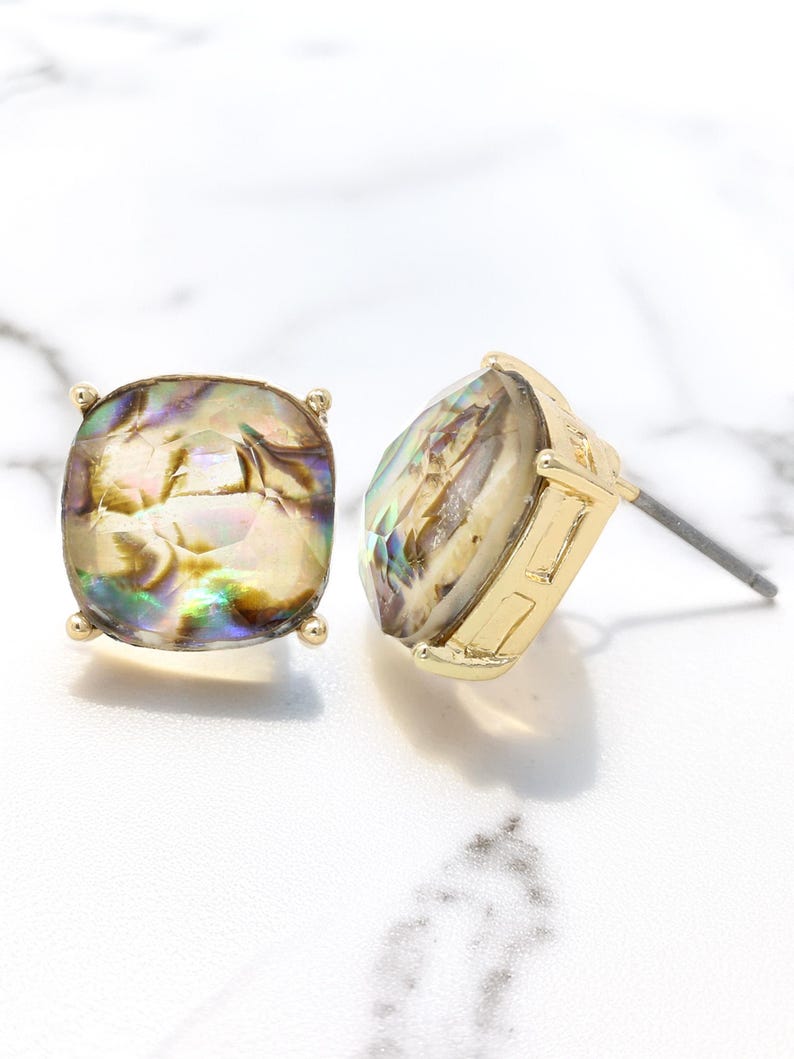 Puede incluir: Dos pendientes de bot&oacute;n dorados con piedras cuadradas iridiscentes. Las piedras muestran una mezcla de colores, incluyendo oro, verde y p&uacute;rpura. Cada pendiente tiene un poste para orejas perforadas y peque&ntilde;os detalles dorados en las esquinas.