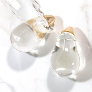 May include: Two clear, teardrop-shaped glass perfume bottles with gold-colored caps. The bottles are filled with a clear liquid and sit on a white marble surface. The gold caps add a touch of elegance to the design.