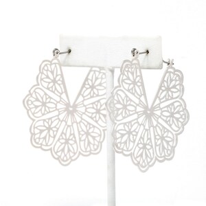 May include: A pair of silver-colored, openwork earrings. Each earring features a fan-like design with intricate floral patterns. The earrings are displayed on a white stand against a white background, showcasing their delicate details.