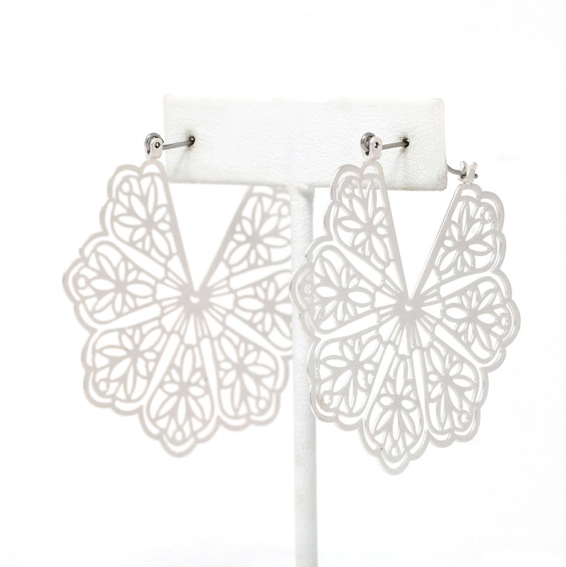 May include: A pair of silver-colored, openwork earrings. Each earring features a fan-like design with intricate floral patterns. The earrings are displayed on a white stand against a white background, showcasing their delicate details.
