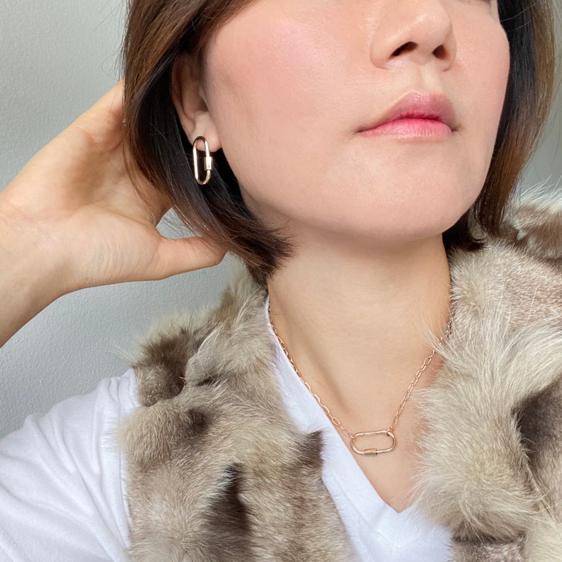 May include: A gold chain necklace with a gold oval-shaped pendant. The pendant is a simple, minimalist design. The necklace is worn by a person with short brown hair and a fur vest.