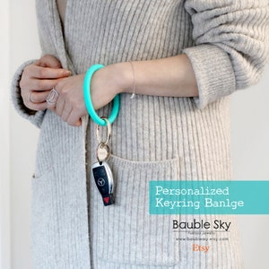 May include: A turquoise silicone keyring bangle with a gold key ring and a silver car key. The text "Personalized Keyring Bangle" is displayed in white on a turquoise background. The text "Bauble Sky" is displayed in white on a turquoise background. The text "Fashion Jewelry" is displayed in white on a turquoise background. The text "www.baublesky.etsy.com" is displayed in white on a turquoise background. The text "Etsy" is displayed in white on a turquoise background.