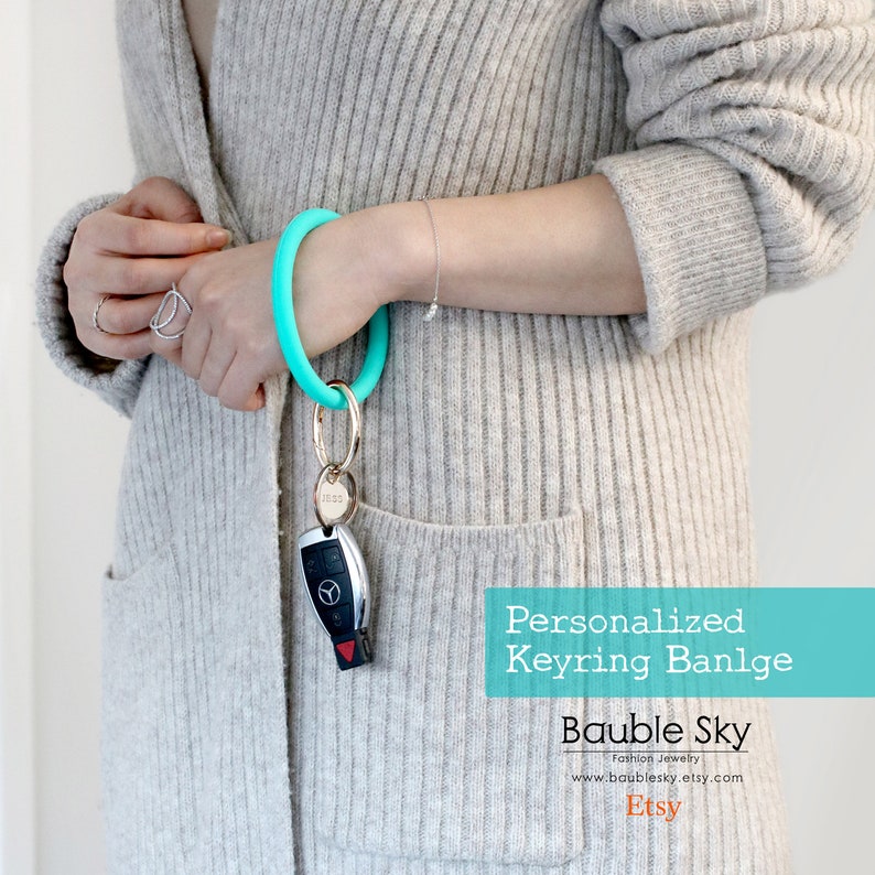 May include: A turquoise silicone keyring bangle with a gold key ring and a silver car key. The text "Personalized Keyring Bangle" is displayed in white on a turquoise background. The text "Bauble Sky" is displayed in white on a turquoise background. The text "Fashion Jewelry" is displayed in white on a turquoise background. The text "www.baublesky.etsy.com" is displayed in white on a turquoise background. The text "Etsy" is displayed in white on a turquoise background.