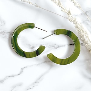 May include: A pair of olive green hoop earrings with a translucent, marbled texture and silver-toned metal posts. The earrings feature a circular, open-hoop design with a smooth, polished finish that catches the light, showcasing deep green swirls.
