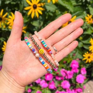 May include: Three bracelets stacked on a wrist. The bottom bracelet is a multi-colored striped beaded bracelet. The middle bracelet is a gold beaded bracelet with a white pearl and a yellow smiley face charm. The top bracelet is a white beaded bracelet.